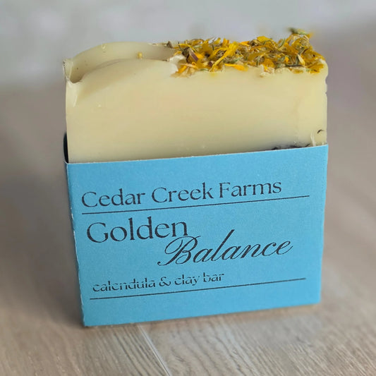 Cedar Creek Farms Golden Balance Botanical Bar front label close-up showing calendula and clay soap bar with blue label on wooden surface